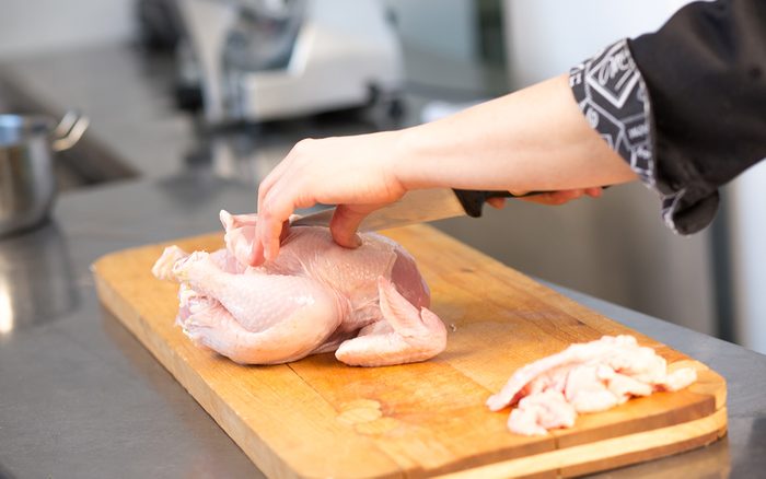 A woman chef caucasian cuts raw chicken. Master class. Cook's hand with a knife close-up on the background of the kitchen. The background is blurred