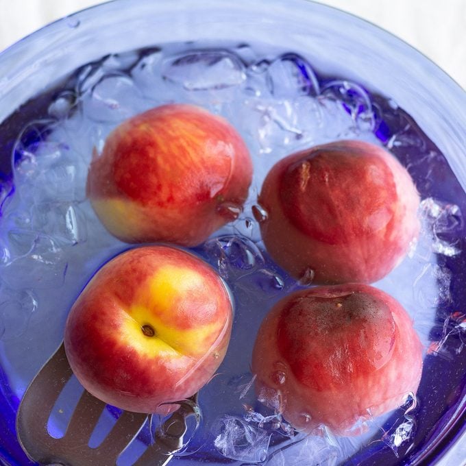 Blanched whole peaches in a blue bowl of ice water.
