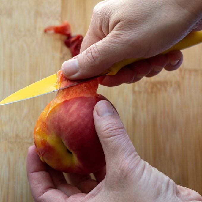Using a yellow paring knife to peel skin from a blanched whole peach.