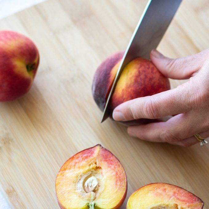 Slicing a whole peach in half on a cutting board with a cut peach nearby.