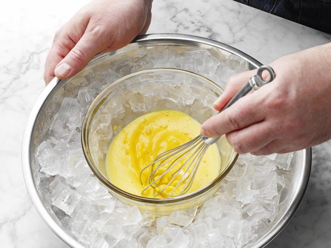 hands whisking eggs in a glass bowl that rests in a larger bowl filled with ice