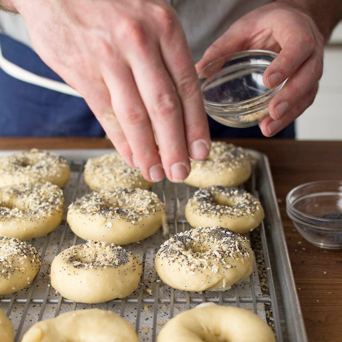 A person seasoning a tray of homemade boiled bagels before baking them.