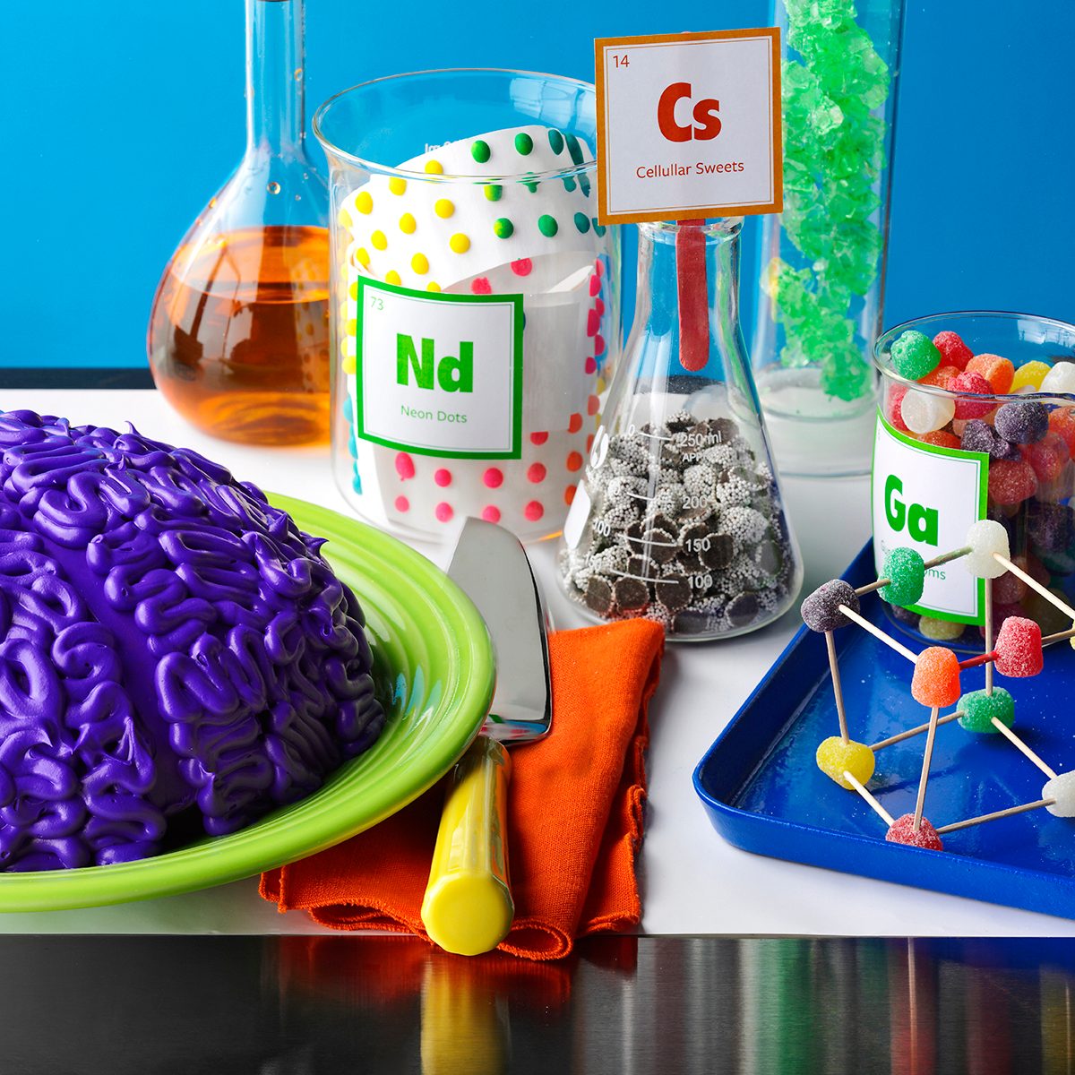 Purple 'brain' on a green plate surrounded by beakers and jars of labelled candy and juice