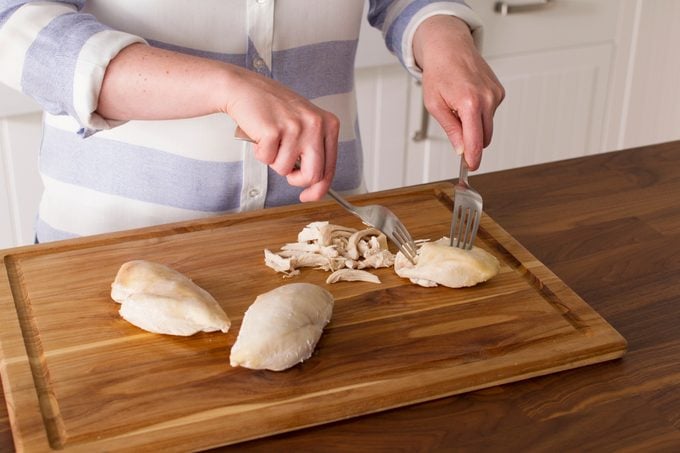Person using two forks to shred chicken breasts on a wooden cutting board