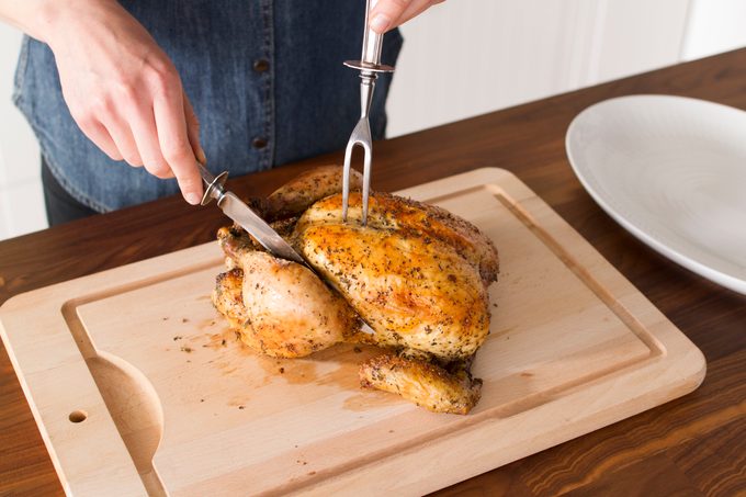 Person holding a whole cooked chicken down with a fork as they slice into one leg with a knife