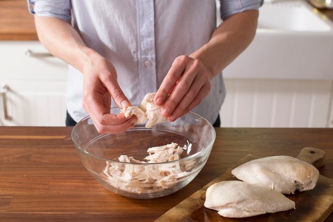 Person using their hands to shred chicken meat over a glass bowl