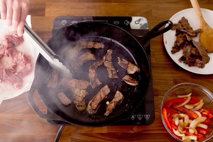 Person stirring sliced meat in a cast-iron skillet with metal thongs