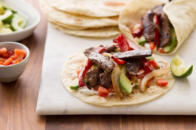 Two fajitas beside a stack of tortillas and bowls of veggies