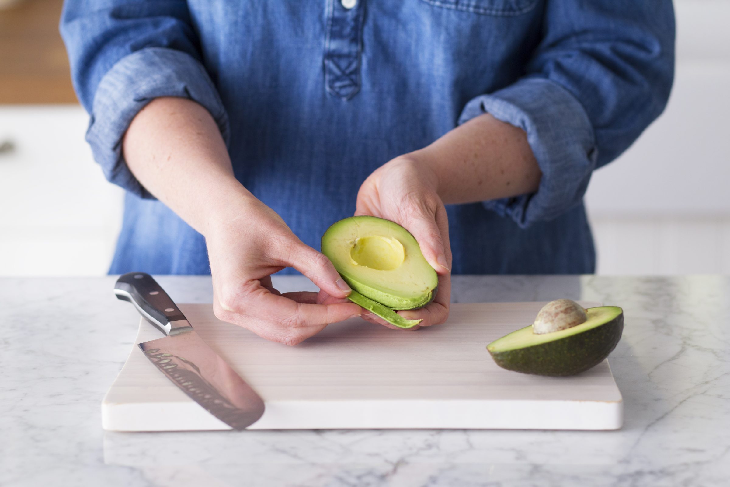 Person peeling the skin off an avocado that is sliced in half over a cutting board