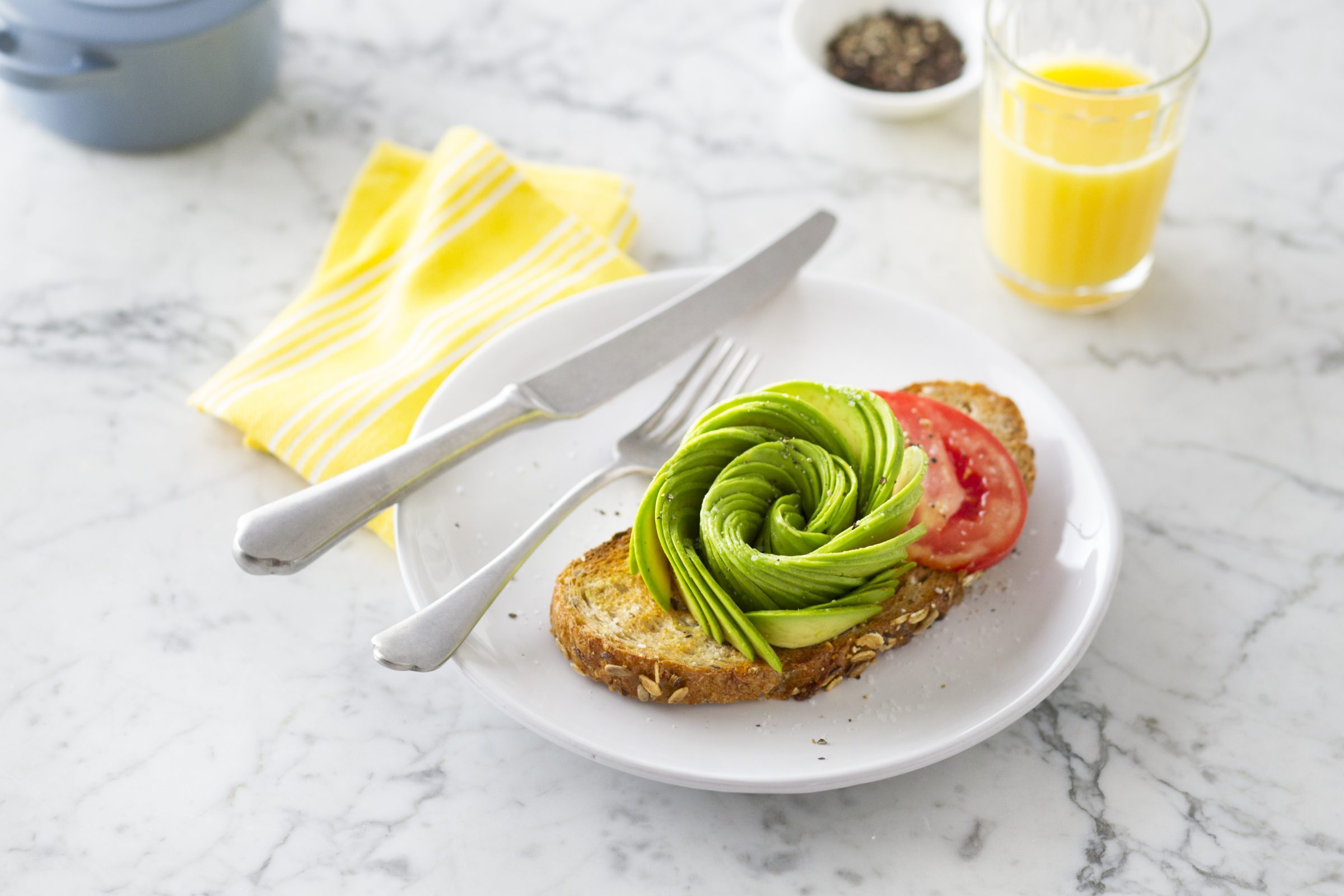 Avocado rose on top of a slice of toast on a plate beside a knife and yellow napkin on marble countertop