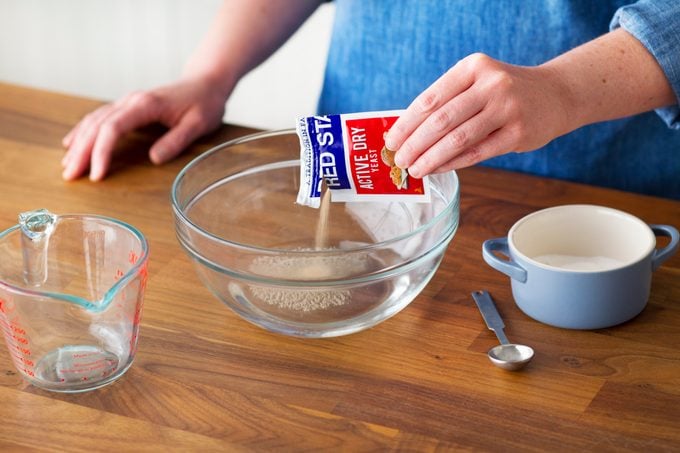 Person pouring an open packet of yeast into sugar and water inside a glass bowl