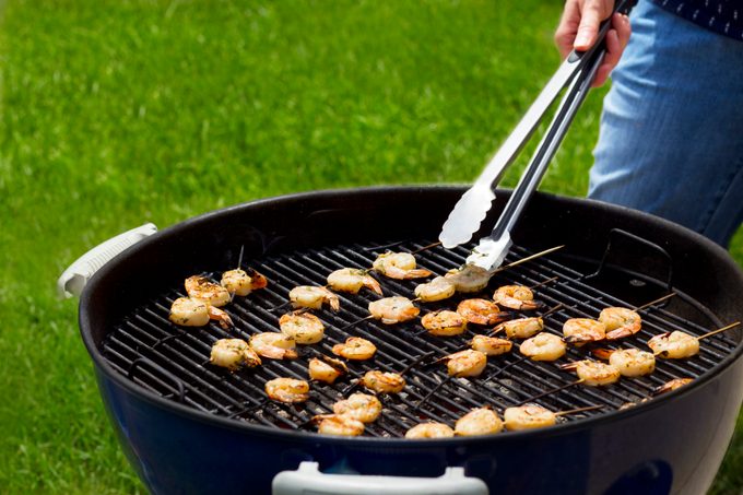 Person using metal tongs to flip skewered shrimp on a charcoal grill