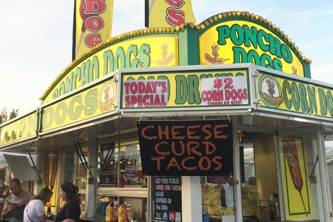 Stand at a state fair with bright yellow signs announcing "Poncho Dogs" and a chalk board advertising cheese curd tacos