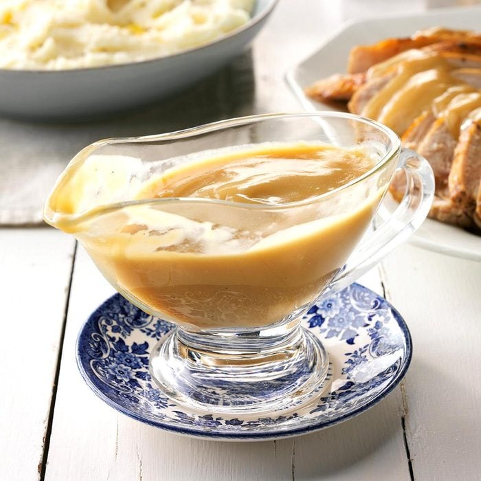 Make-Ahead Turkey Gravy in glass gravy boat on a saucer on a white table. other thanskgiving food in the background