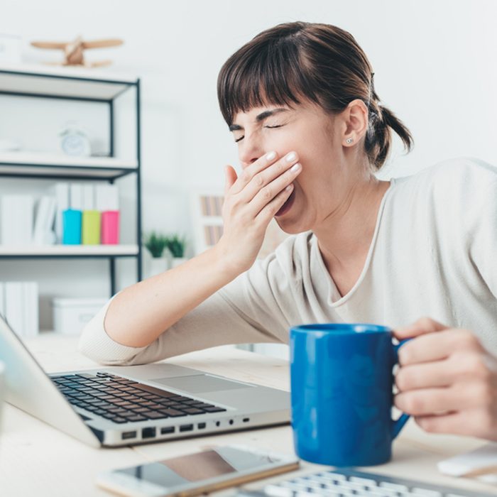 Tired sleepy woman yawning, working at office desk and holding a cup of coffee, overwork and sleep deprivation concept; Shutterstock ID 367741853
