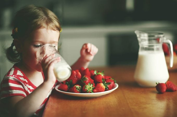 Happy child girl drinks milk and eats strawberries in the summer home kitchen