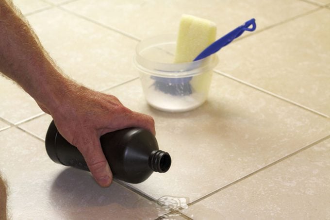 Hand pouring hydrogen peroxide on bathroom floor tile grout.