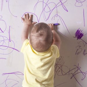 Baby boy drawing with wax crayon on plasterboard wall. He is with his back towards