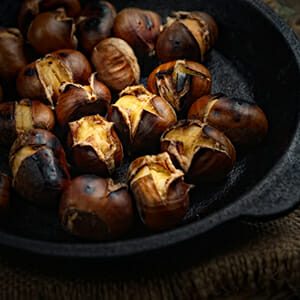 Heap of grilled edible chestnuts in cast iron skillet over dark wooden surface with textile napkin