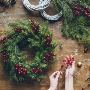 top view of florist hands making Christmas wreath with fir branches and decorative berries on wooden tabletop