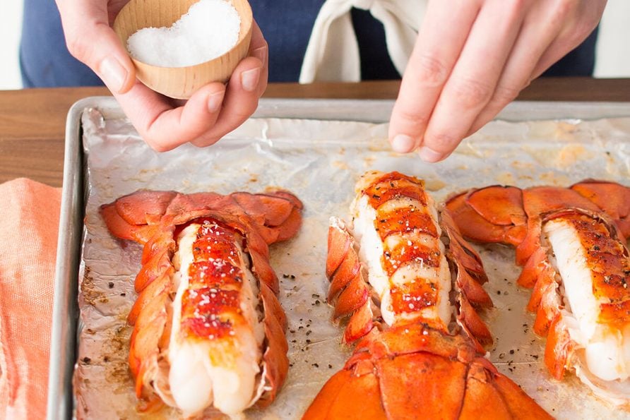 Person sprinkling salt over lobster tails on a baking sheet