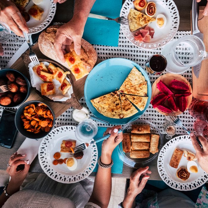 Above aerial view of group of friends having fun eating together at lunch or dinner with a table full of different and colorful food and technology mobile phone. mix of hands of caucasian people ; Shutterstock ID 1196986168; Job (TFH, TOH, RD, BNB, CWM, CM): TOH