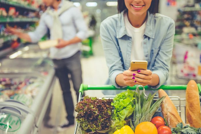 Cropped image of girl leaning on shopping cart, using a mobile phone and smiling, in the background her boyfriend is choosing food