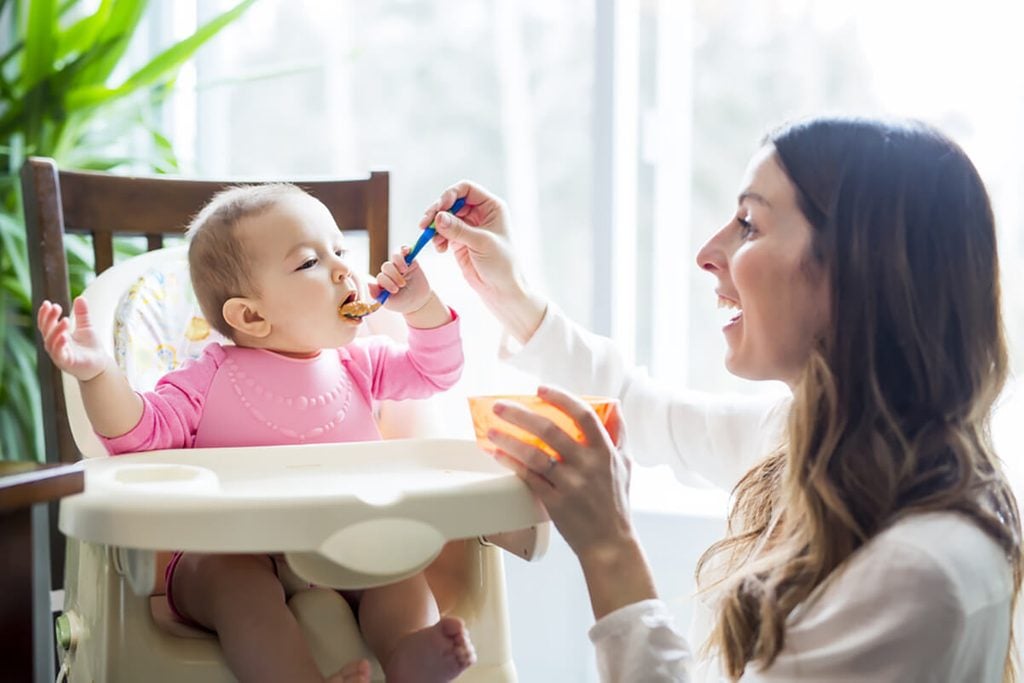 Young Mom Feeding Homemade Baby Food to Daughter in High Chair