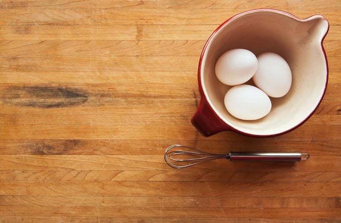 A view looking down on a red ceramic bowl filled with eggs on a butcher block counter with a whisk sitting to the side