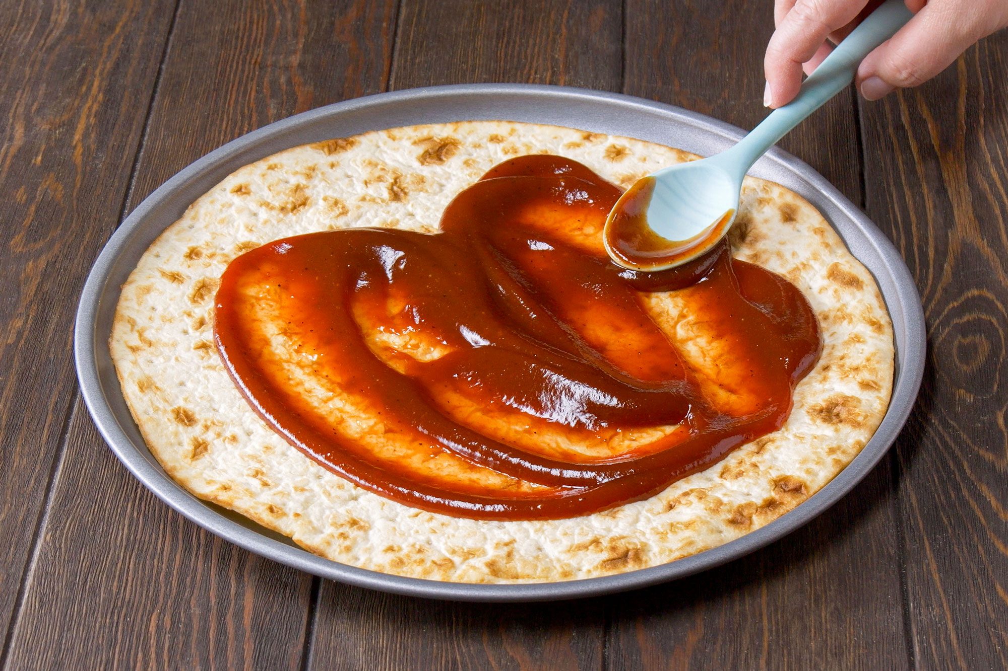 Applying Barbecue Sauce on Pizza Crust with a Plastic Spoon, Wooden Background