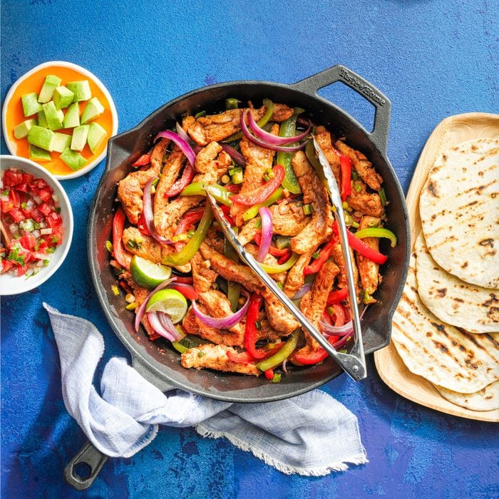 Chicken Fajitas in a Large Cast Iron Skillet and Tortillas in a Wooden Plate on Blue Background