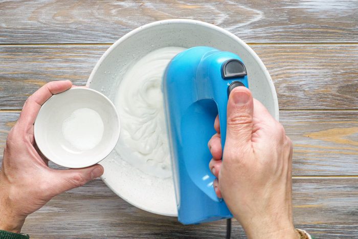 Beating Egg Whites in a White Large Bowl with Hand Mixer. Sugar in a Small Bowl in Left Hand