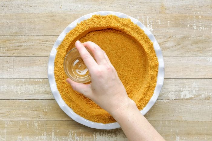 Pressing Graham Cracker Crumbs into the Deep-Dish Pie Plate with the help of Glass on Wooden Surface to make Easy Cream Pie