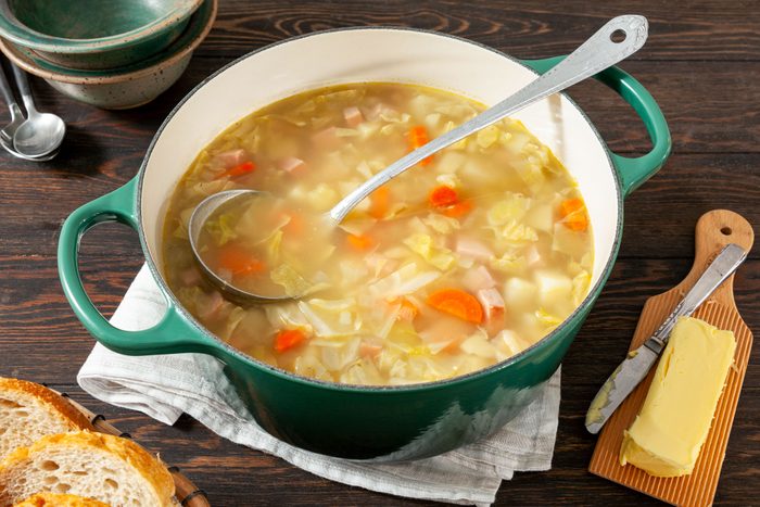 Ham And Cabbage Soup in Ceramic Soup Pot on Wooden Surface with Butter on Right Lower Corner and Empty Bowls on Left Upper Corner