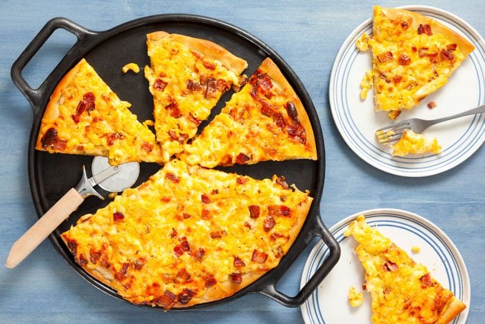 Mac And Cheese Pizza on a Cast Iron Pizza Skillet and Pizza Slices Served on White Ceramic Plates on Textured Blue Background