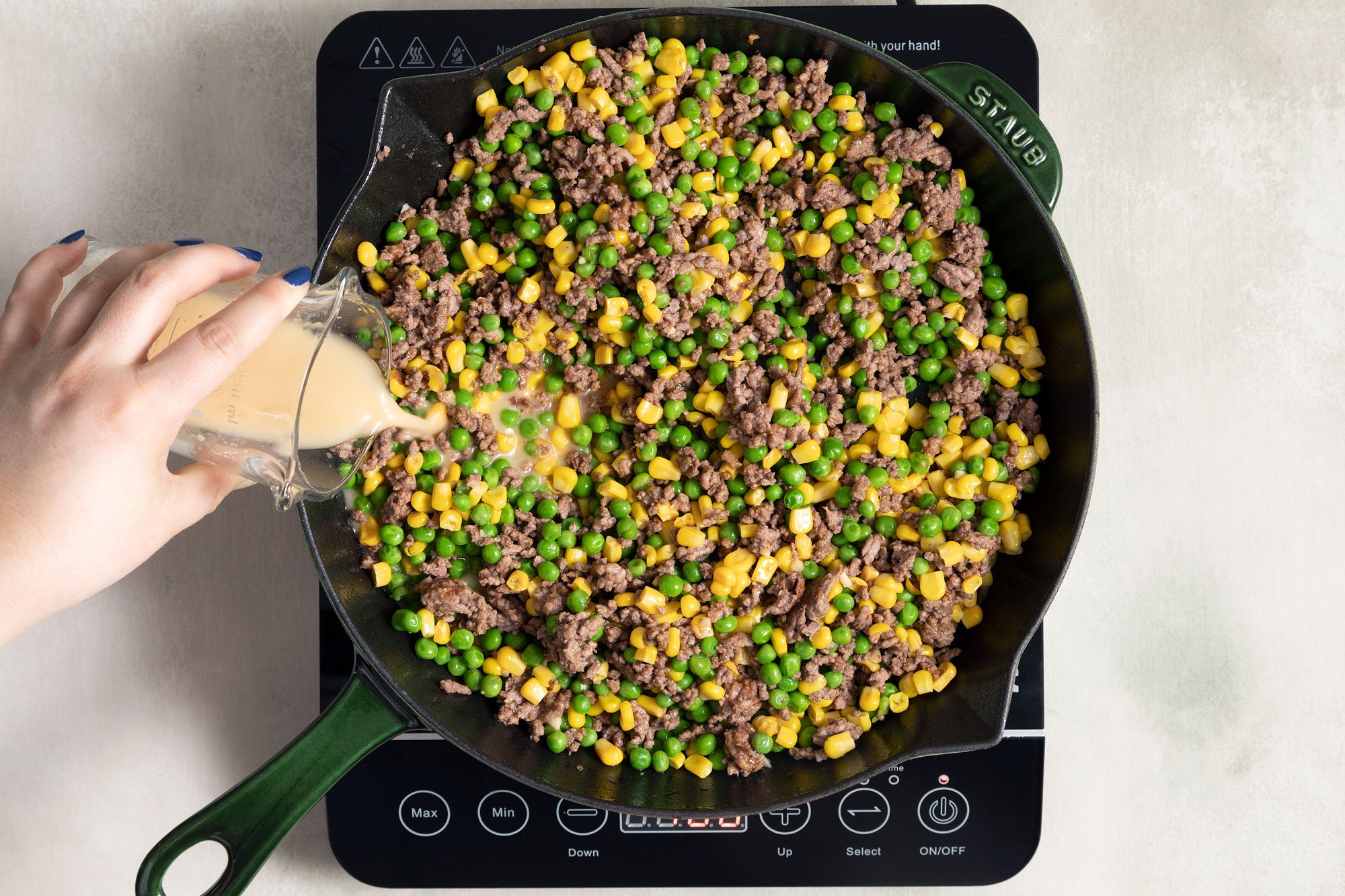 someone pouring cornstarch-water mix into the mixture of cooked beef, corn, peas in a large skillet on induction cooktop