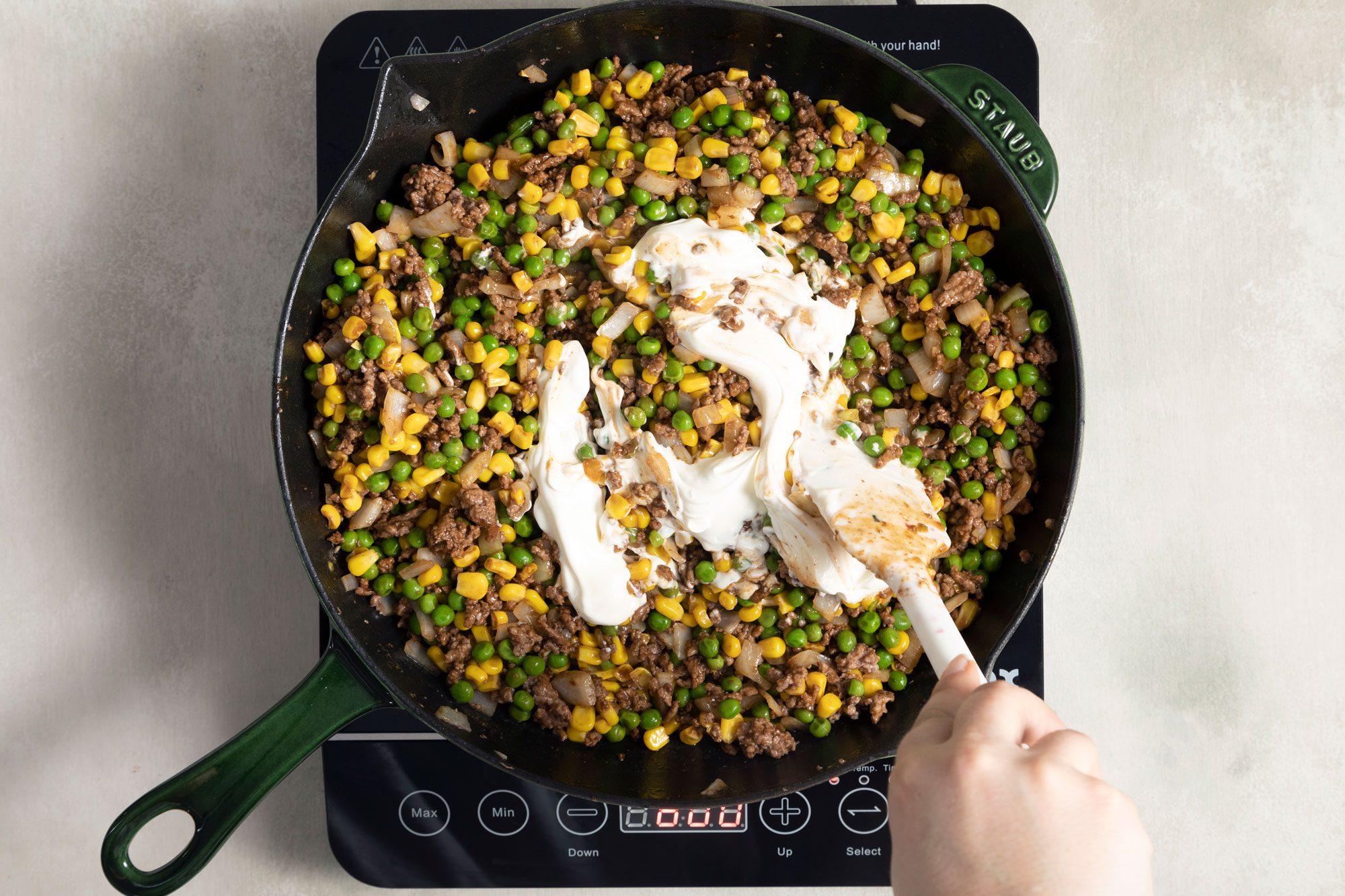 someone mixing sour cream into the mixture of cooked beef, corn, peas in a large skillet on induction cooktop