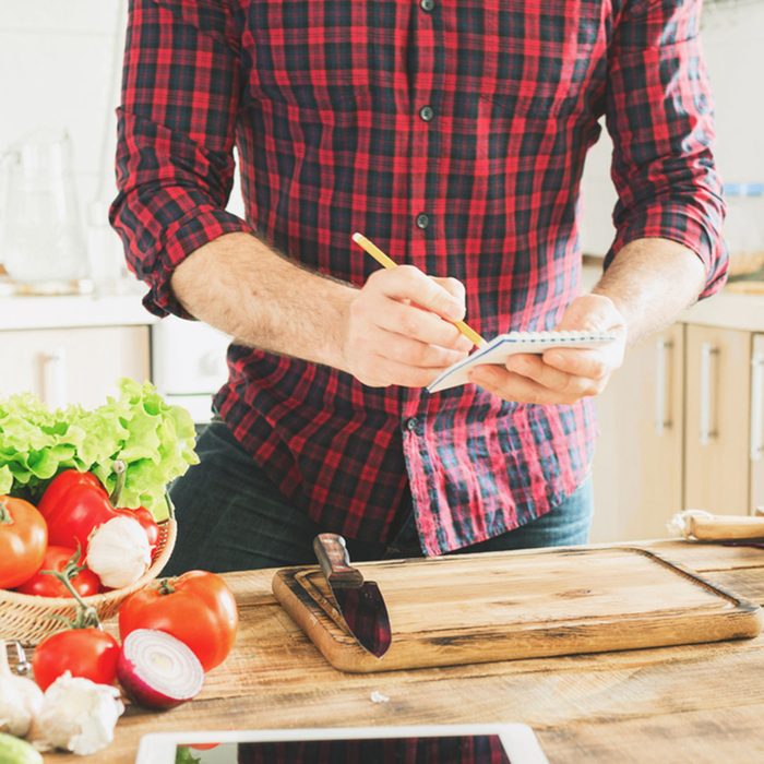 Ingredients for cooking healthy food on a wooden table in a home kitchen. In the background a man writes a recipe to notebook; Shutterstock ID 556856305