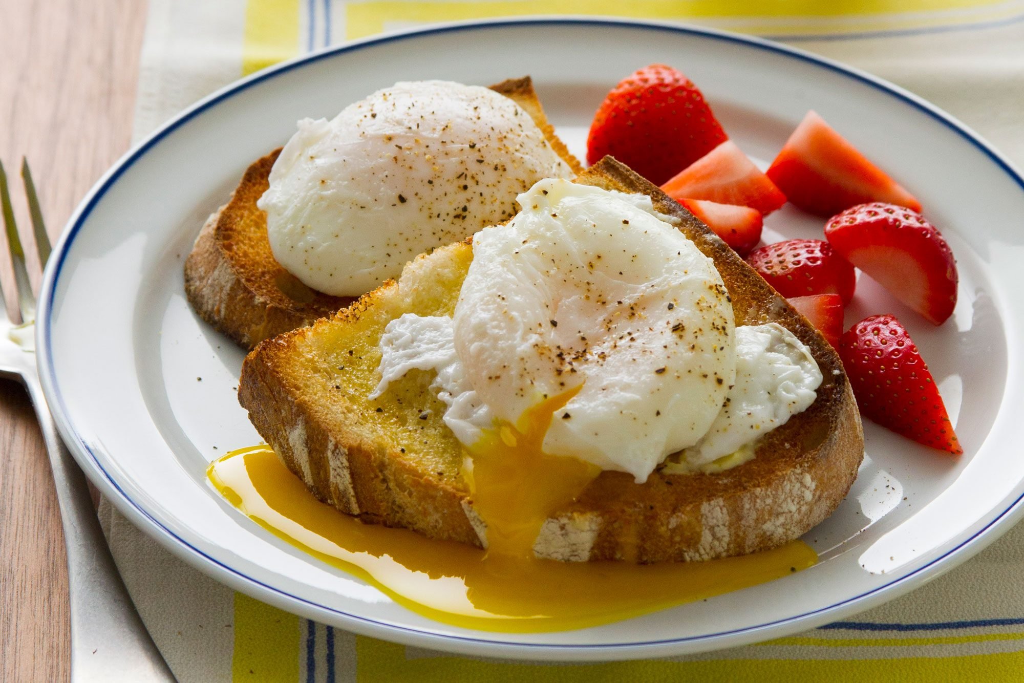 poached eggs on toast and strawberries on a white plate on a table next to a fork. the yolk from one egg has been broken and is dripping onto the toast and plate