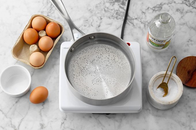 pan with water on a hot plate surrounded by other eggs and other supplies for making poached eggs