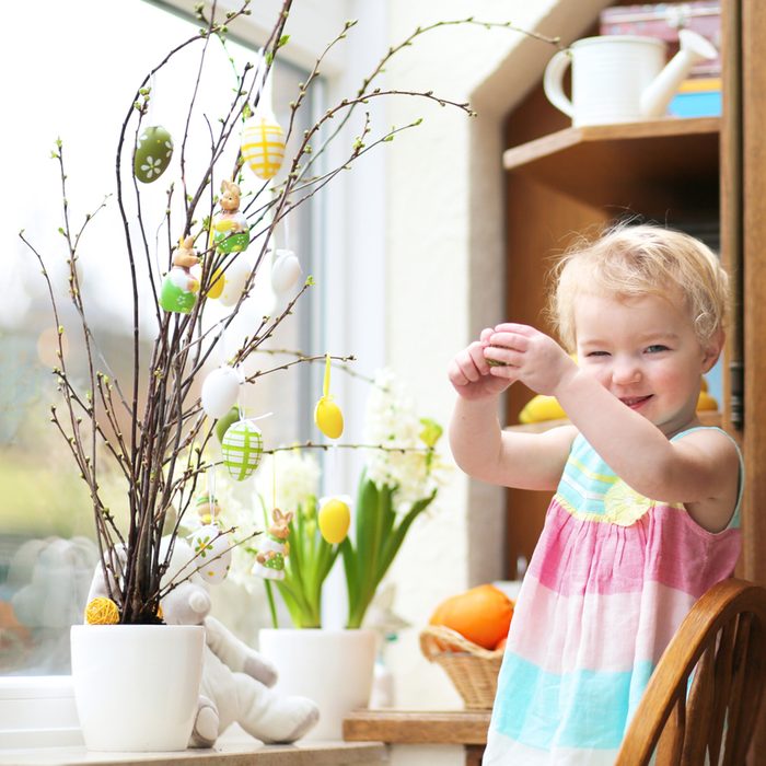 Adorable little blonde toddler girl decorating with Easter eggs cherry tree branches standing in the kitchen next a window with garden view; Shutterstock ID 179985827