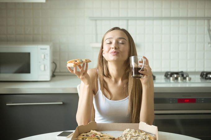 Portrait of a smiling woman holding a slice of pizza in her hand, her eyes closed with pleasure, sitting at the kitchen table, enjoying the taste of pizza.