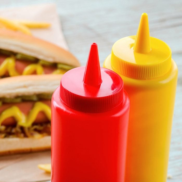 Ketchup and mustard bottles, close-up. Traditional hot dogs and potatoes on the background, baking paper. Western diet