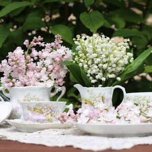 Lilac, bunch, bouquet of lily of the valley in vintage, antique authentic china, porcelain milk jug, sugar bowl, tea cup with saucer on lace napkin on table, bush background, garden floral scene