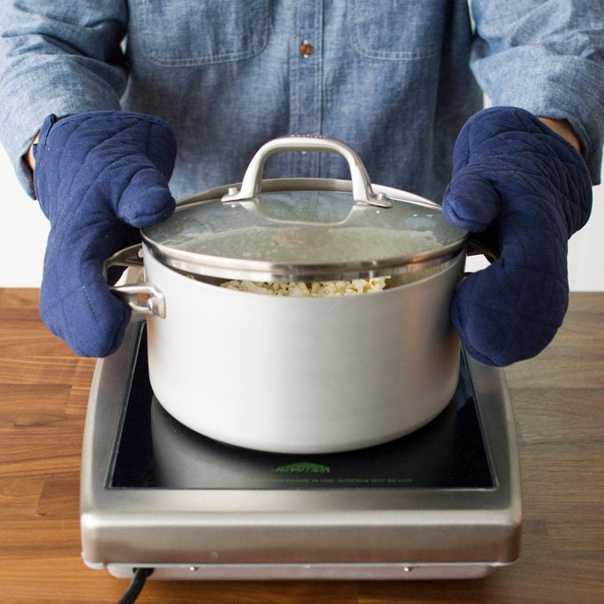 A person wearing oven mitts and shaking a pot of homemade stovetop popcorn over an electric burner.