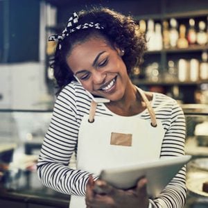 Smiling young black entrepreneur standing at the counter of her cafe talking on a cellphone and using a tablet