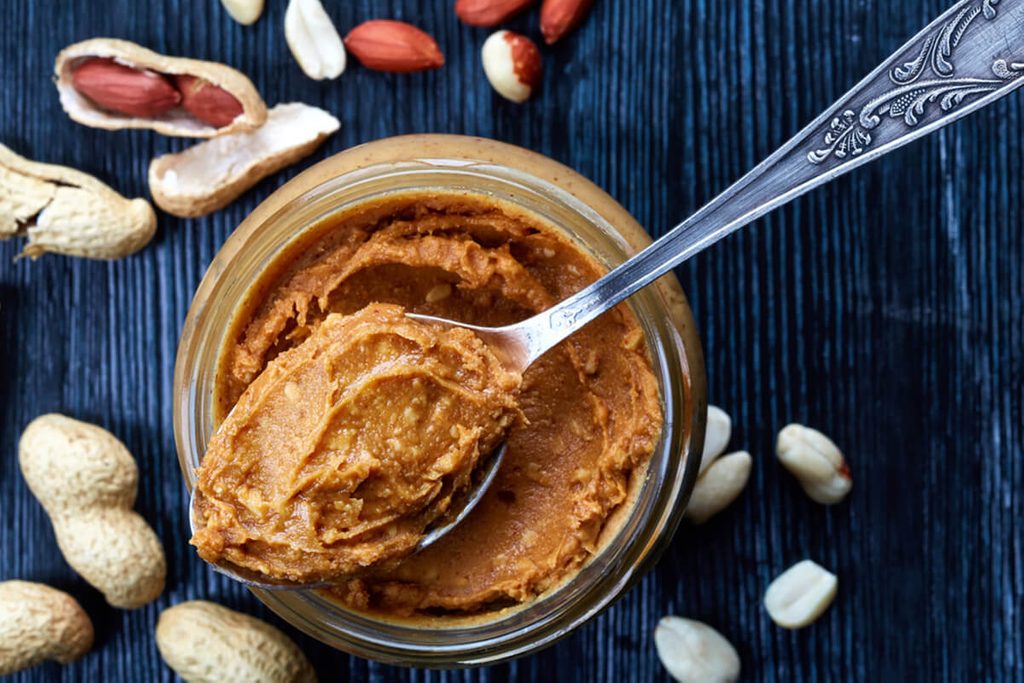 Jar and spoon of peanut butter and peanuts on dark wooden background from top view