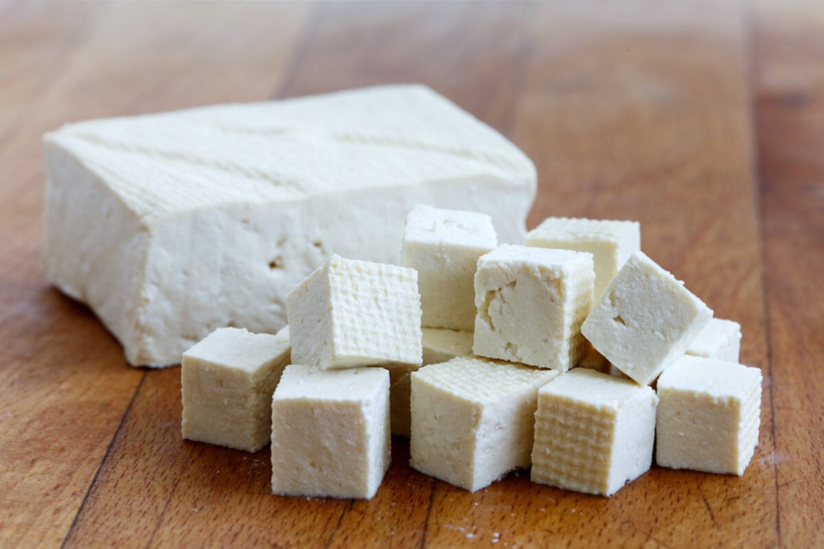 Single block of white tofu with cut tofu cubes on wooden chopping board