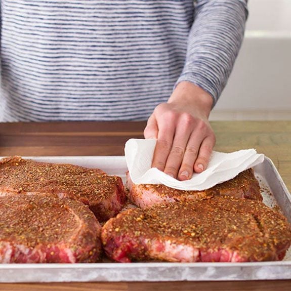 Patting seasoned raw steaks with a paper towel