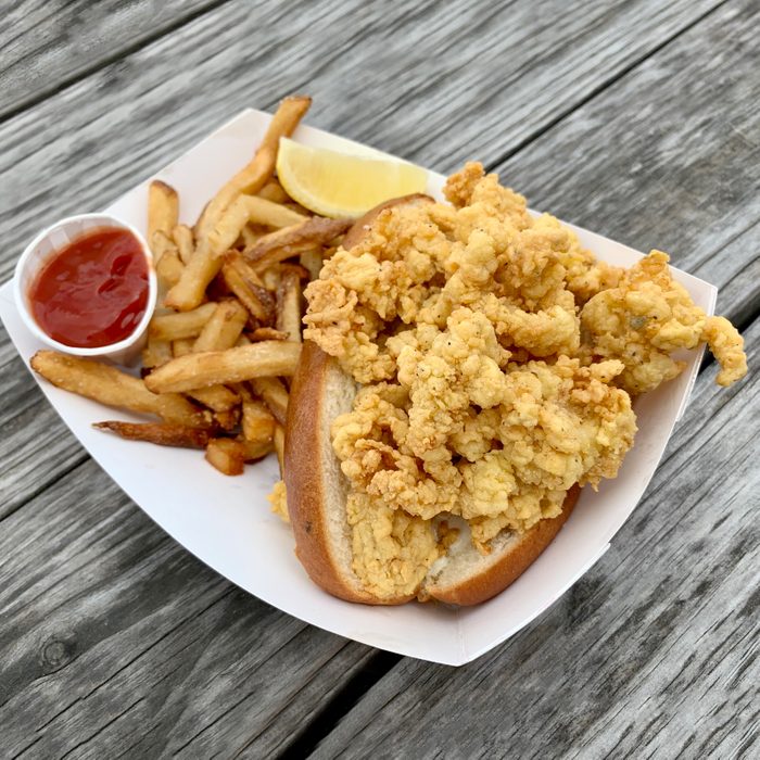 Fried Clam Roll Sandwich with fries and ketchup on a white plate resting on a wooden surface